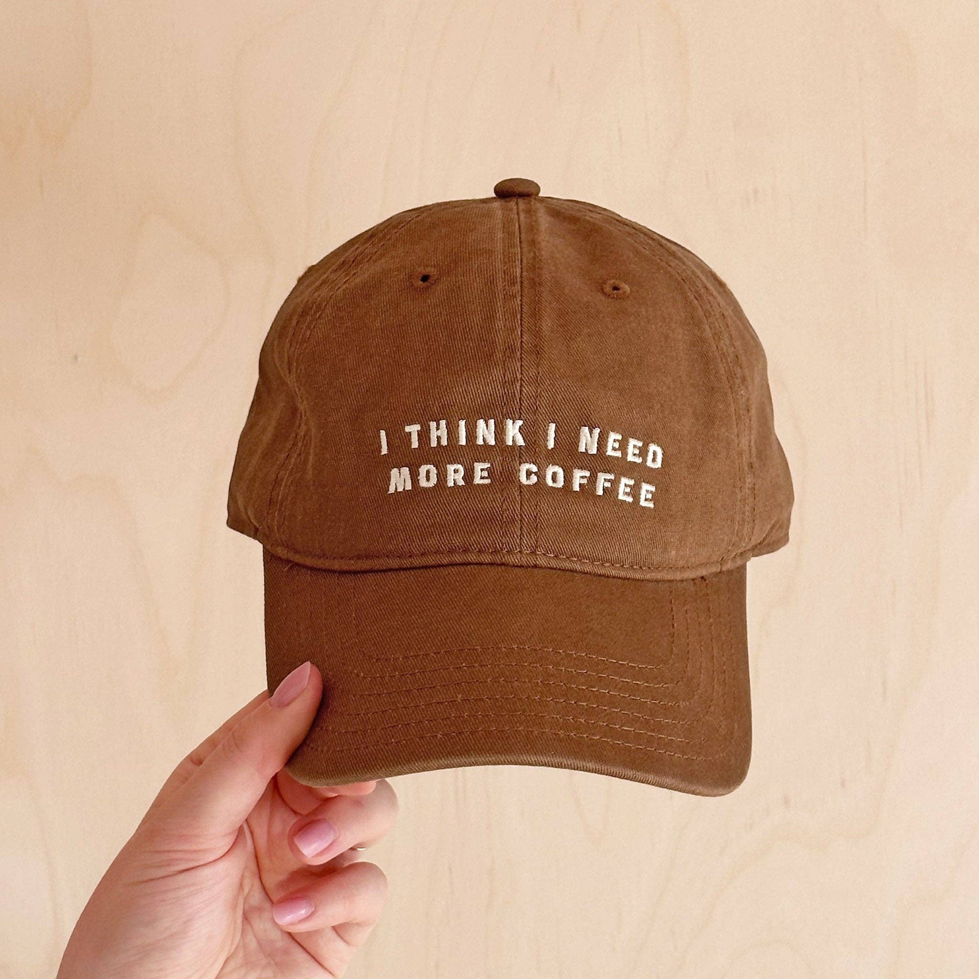 Brown cap with 'I think I need more coffee' text held by a hand on a beige background