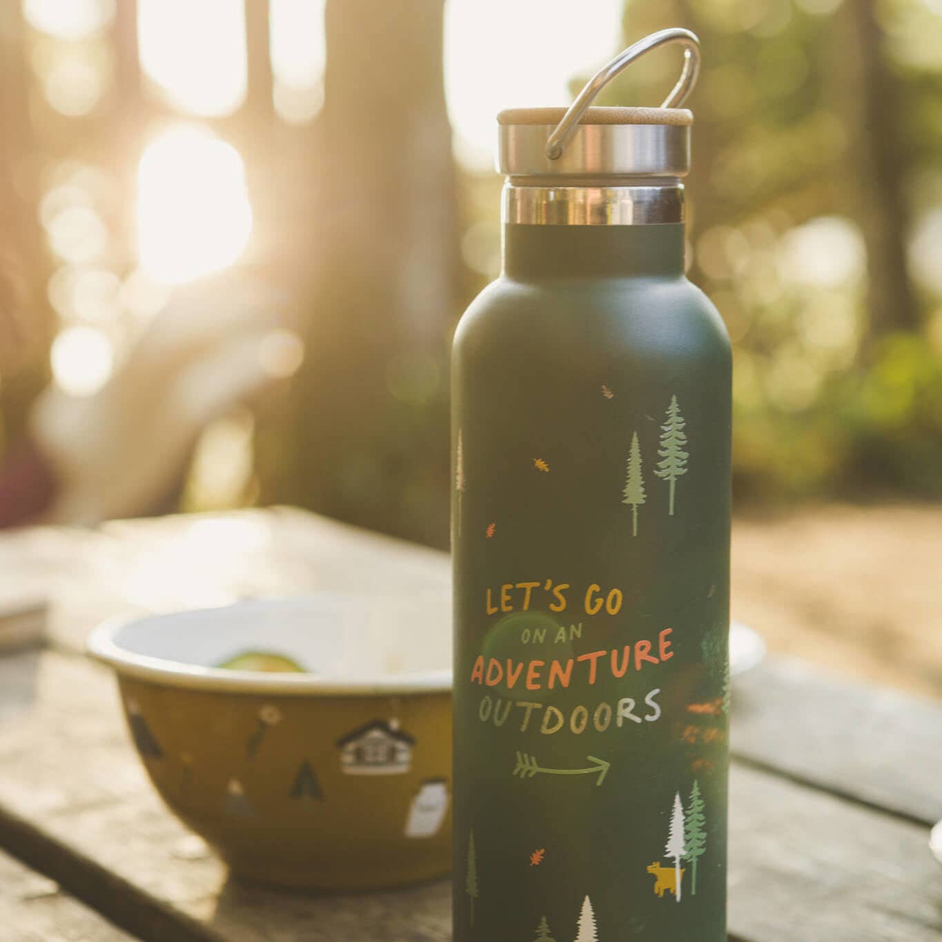 Green water bottle with 'Let's go on an adventure outdoors' text and nature-themed design on a wooden table with a blurred forest background.